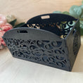 Load image into Gallery viewer, A black laser-cut wooden gift basket with decorative scrollwork patterns and two handles, shown on a light wood surface with pink flowers and green leaves in the background.