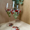 Load image into Gallery viewer, Two clear wine glasses decorated with hand-painted red flowers and green leaves, resting on a light-colored wooden surface with a blurred background.