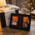 Load image into Gallery viewer, A gift set displayed on a wooden table, featuring a black presentation box with two floral-themed items and a black pen. The scene is styled with a warm lamp, a knitted blanket, a steaming mug, and fairy lights in the background.