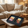 Load image into Gallery viewer, A black gift box set displayed on a rustic wooden table in a cozy living room. The set includes two printed cards or magnets with a red poppy field landscape design and a black pen.