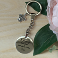 Load image into Gallery viewer, A silver metal keychain with a round engraved disc charm reading 'Be proud of who you have named. Eva', a small sea turtle charm, and a split key ring, styled on a light wood surface with green leaves and a pink flower.
