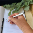 Load image into Gallery viewer, A close-up of a hand holding a white pen with a black tip, writing in a lined notebook. A second black pen rests on the notebook, and blurred green eucalyptus leaves are in the background.