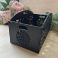 Load image into Gallery viewer, A black wooden decorative gift basket with laser-cut circular patterns and side handles, shown on a light beige tabletop with soft pink flowers in the background.