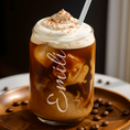 Load image into Gallery viewer, A frosted glass iced coffee drink topped with whipped cream and cocoa powder, with the name 'Emili' written in white script on the glass, set on a round wooden tray with scattered coffee beans.