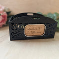 Load image into Gallery viewer, A black laser-cut decorative tray with a central wooden plaque engraved with the name and Happy 50th Birthday', set on a light wooden surface with blurred pink flowers and green leaves in the background.
