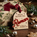 Load image into Gallery viewer, A personalized Christmas gift tag reading 'SPECIAL DELIVERY FOR: Roman TO OPEN ON DECEMBER 25TH' in red lettering, attached to a wrapped gift with holly and pine illustrations and a dark red ribbon, styled on a wooden surface with holiday greenery, ornaments, and gingerbread cookies.
