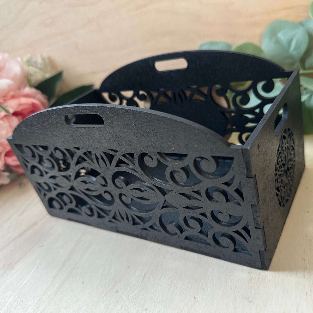 A black laser-cut wooden gift basket with decorative scrollwork patterns and two handles, shown on a light wood surface with pink flowers and green leaves in the background.