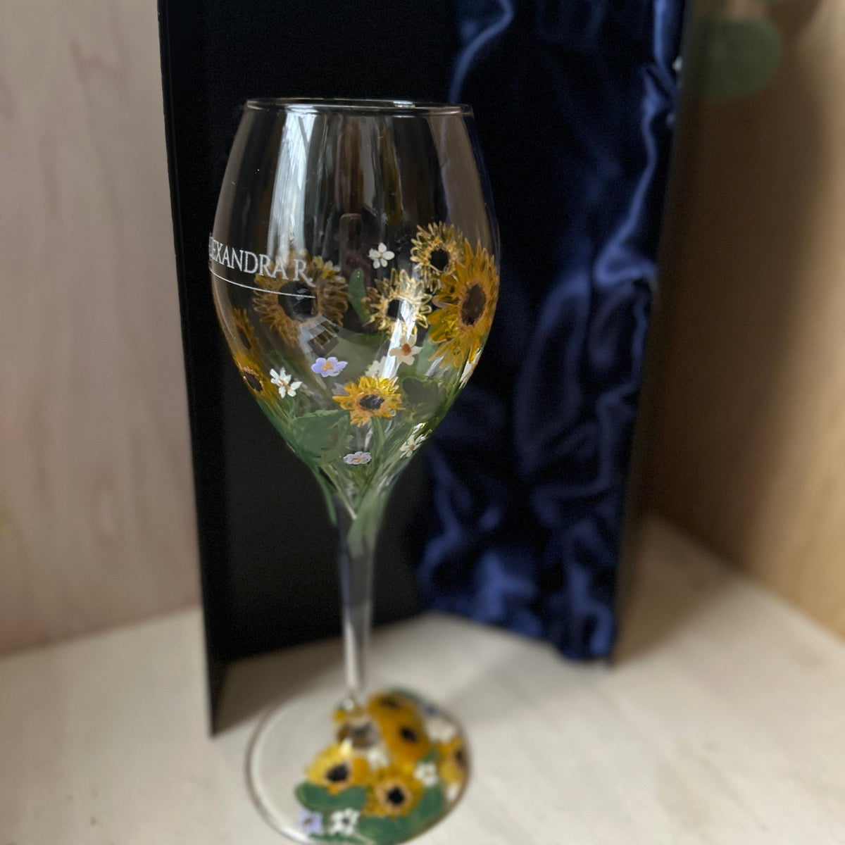 A clear wine glass decorated with yellow sunflowers and small white flowers, shown in front of a black presentation box with dark blue satin lining on a light wood surface.