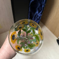 Load image into Gallery viewer, A hand holds a clear glass orb decorated with pressed sunflowers, green leaves, and small white flowers. A black presentation box with a blue satin lining is standing behind it on a light-colored surface.
