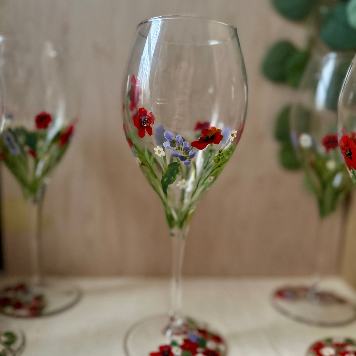 A clear wine glass with hand-painted red, purple, and green floral designs near the bowl and base, displayed on a light tabletop with other similar glasses blurred in the background.