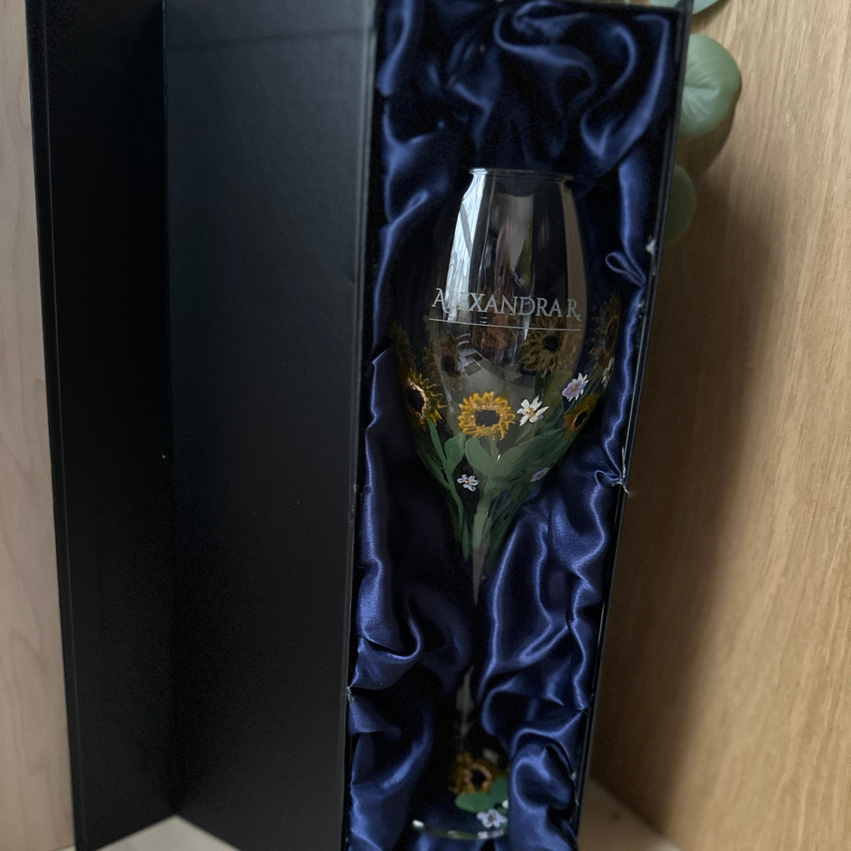 A hand-painted glass wine glass with floral designs and the name printed on it, presented inside an open black gift box lined with dark blue satin.