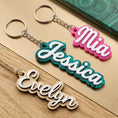 Load image into Gallery viewer, Three personalized name keychains spelling 'Mia', 'Jessica', and 'Evelyn' in script lettering, shown on a light wood background with a green patterned card partially visible at the top right.
