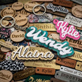 Load image into Gallery viewer, Three custom wooden name keychains with silver keyrings are displayed on top of a pile of other engraved wooden name tags. The main keychains feature layered wooden letters with different colored felt trim: one with teal felt trim and white lettering, one with pink felt trim and white lettering, and one with beige felt trim and white lettering.
