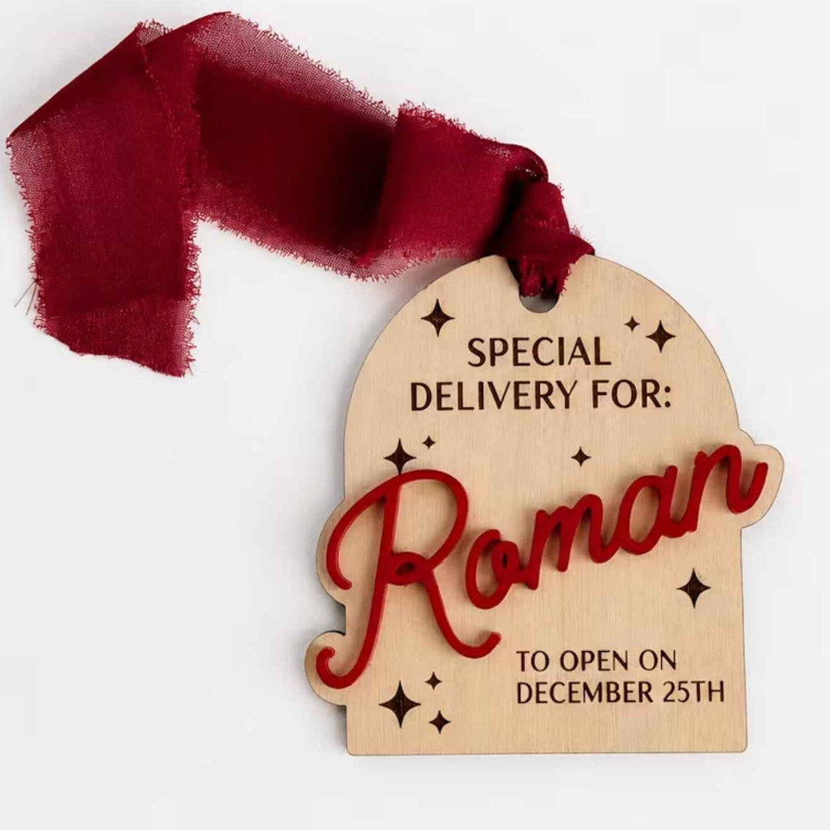 A personalized wooden arch-shaped gift tag with 3d name hanging with a burgundy ribbon loop, featuring engraved text that reads 'SPECIAL DELIVERY FOR: Roman TO OPEN ON DECEMBER 25TH' with decorative star details.