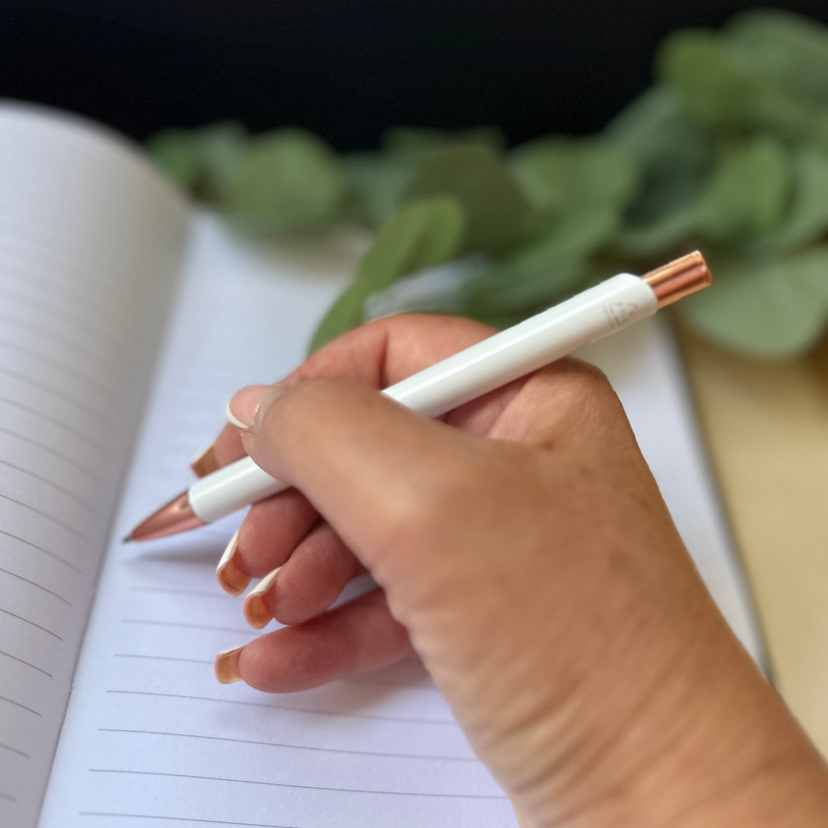 A close-up of a hand holding a white pen with rose gold accents, writing in an open lined notebook with blurred green leaves in the background.