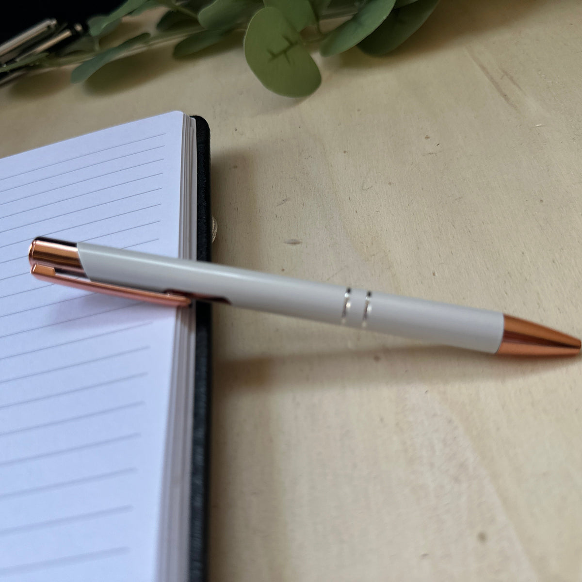 A white ballpoint pen with rose gold accents resting on an open lined notebook on a light wood desk.