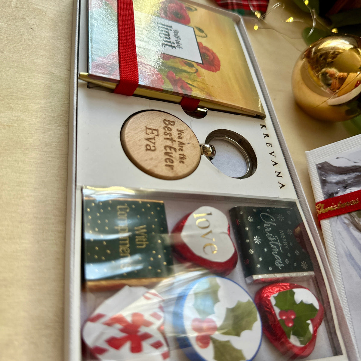 A Christmas-themed gift set in a white box containing a small notebook tied with a red ribbon, a round wooden keychain and 6 small chocolate treats decorated with holiday motifs like holly and candy canes.