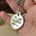 Load image into Gallery viewer, A close-up of a hand holding a silver-toned oval pendant necklace engraved with 'You + Me John & Emma 09.10.
