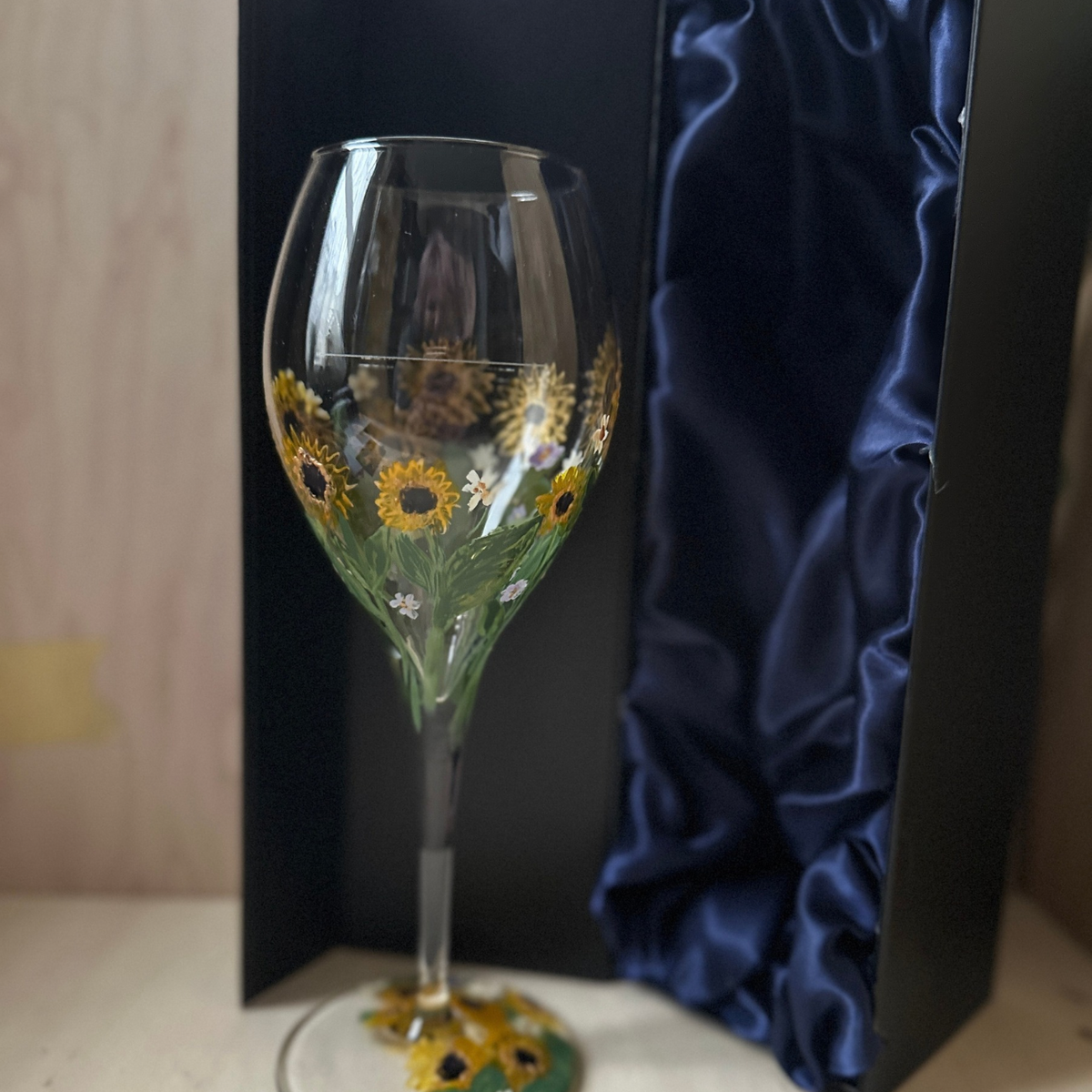 A clear wine glass decorated with painted yellow sunflowers, green stems, and small white flowers, standing in front of a black gift box with a blue satin lining on a light wood surface.