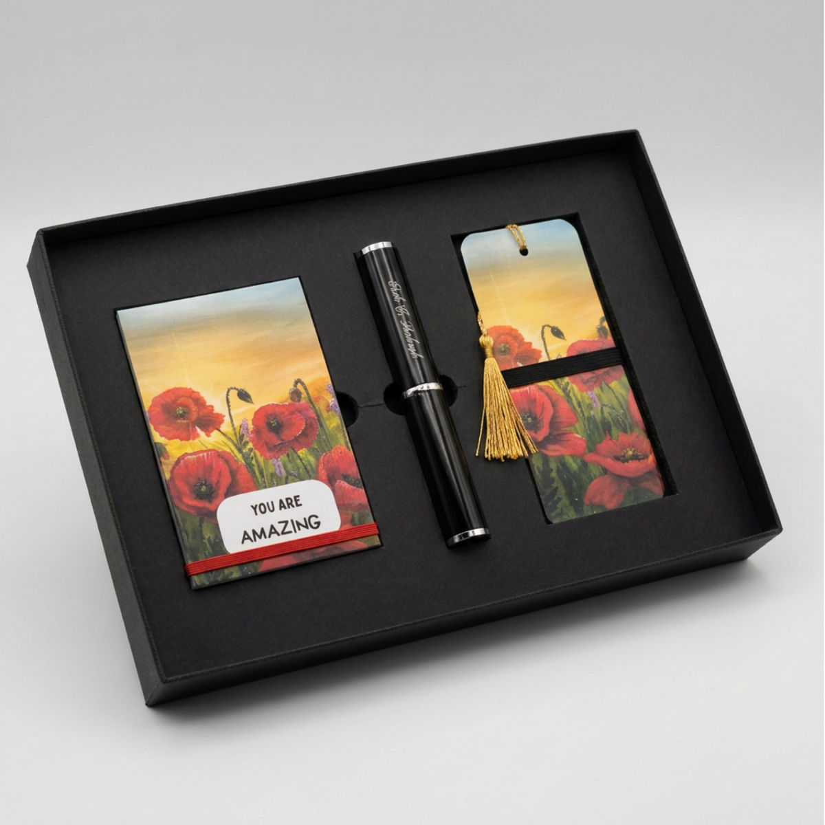A black gift set box containing a black pen and two matching poppy-themed cards or bookmarks with a sunset field print, shown on a light gray background.
