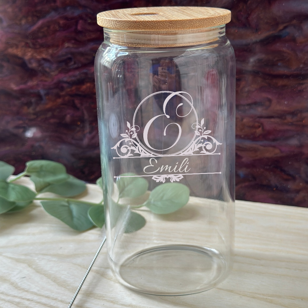 A clear glass jar with a light wood lid, featuring an engraved white monogram and the name 'Emily'. It is placed on a light wooden surface with a metal spoon resting beside it, against a dark, textured background.