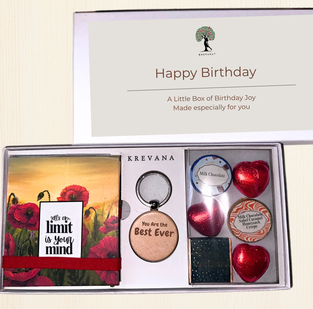 A Krevana Happy Birthday gift box set displayed open on a light beige background. The set includes a red poppy sticky notepad with the quote 'It's your own limit is your mind', a round wooden keychain reading 'You are the Best Ever', milk and dark chocolate hearts, a small tin of gourmet chocolates, 