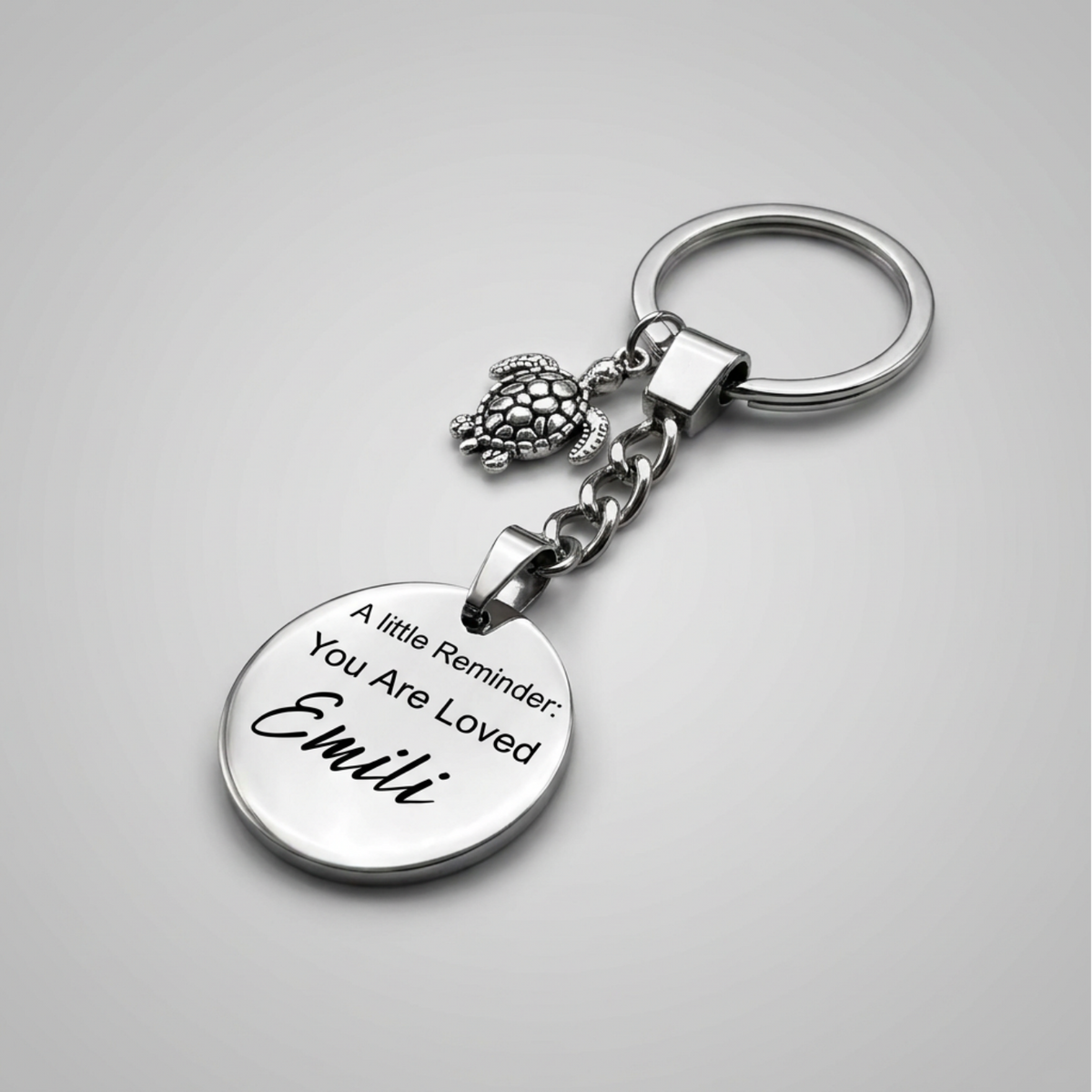 A silver-toned metal keychain with a round engraved disc charm reading 'A little Reminder: You Are Loved Emilé', a small turtle-like charm, and a split key ring, shown on a light gray background.