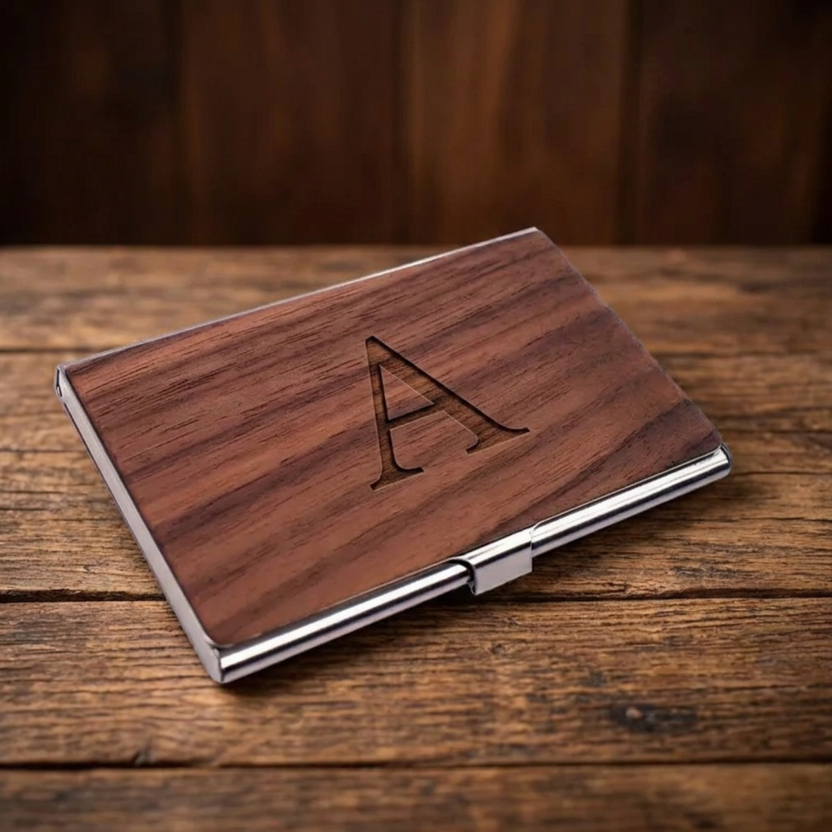 A rectangular wooden business card case with a polished silver metal hinge and border, featuring an engraved capital letter 'A' on the front. The case is displayed on a rustic wooden tabletop with a dark wood background.