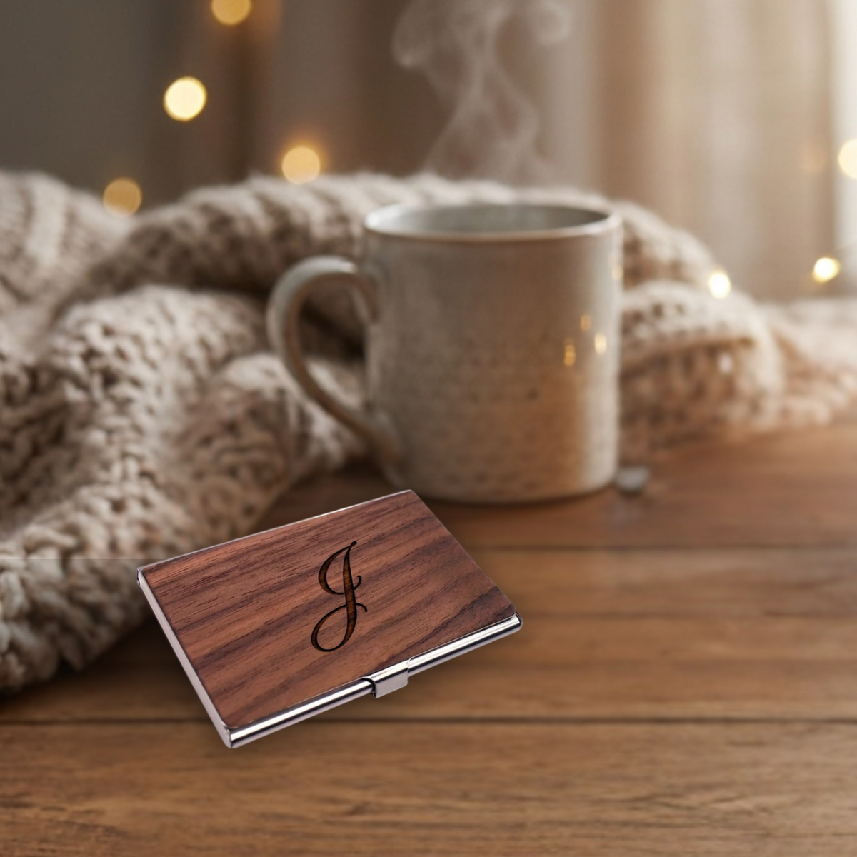 A rectangular wooden card case with a silver metal clasp and an engraved cursive monogram, resting on a wooden table next to a steaming mug and a knitted blanket.