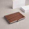Load image into Gallery viewer, A slim rectangular business card case with a brown wood-grain top and polished silver metal edges, photographed on a light beige surface with blurred white cards in the background.
