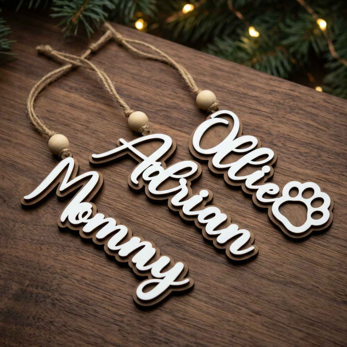 Three personalized wooden hanging ornaments with natural wood edges and string loops, displayed on a dark wood surface with festive greenery and warm string lights in the background. The names read 'Ollie', 'Adrian', and 'Mommy'.