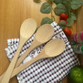 Load image into Gallery viewer, Three light natural wood spoons resting on a white and brown checkered kitchen towel, arranged on a wooden tabletop with decorative green leaves, cherry tomatoes, and a green lime.

