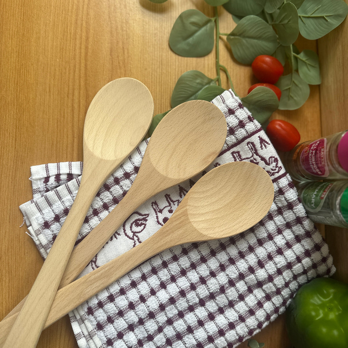 Three light natural wood spoons resting on a white and brown checkered kitchen towel, arranged on a wooden tabletop with decorative green leaves, cherry tomatoes, and a green lime.